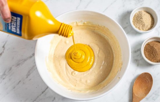 Step 3 — adding mustard and spices to the burger sauce base in a white mixing bowl on a white marble table, horizontal food photography.