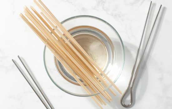 Wooden skewers soaking in water with metal skewers nearby on a white table.