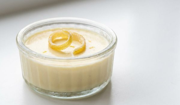 45-degree angle: Professional food photography of a single lemon posset in a glass ramekin, captured from a 45-degree angle, topped with a curl of candied lemon peel. Use soft, bright natural lighting with no harsh shadows and no smoke. Keep the background a clean, uncluttered white surface with subtle negative space. Capture ultra-realistic detail with a shallow depth of field that keeps the ramekin in crisp focus.