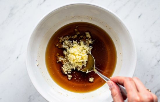 Step 2 — mixing soy sauce, garlic, and ginger together in a white bowl with a small spoon on a white marble surface, bright natural lighting, horizontal format