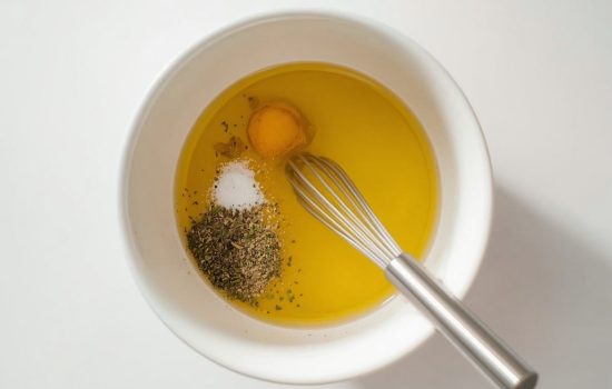 Olive oil, lemon juice, and oregano being whisked in a white ceramic bowl for pasta salad dressing