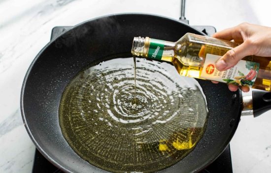 Step 3 — heating vegetable oil in a black non-stick wok on a stove, oil shimmering, hand pouring oil mid-action, bright overhead light on white marble, horizontal format.