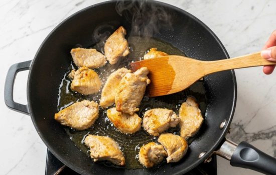 Step 4 — adding raw chicken pieces to a hot black wok with sizzling oil, wooden spatula stirring, chicken turning golden, steam rising, bright overhead light, horizontal format.