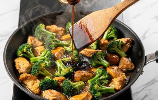 Step 5 — adding bright green broccoli to a black wok with golden chicken, soy sauce being poured over, wooden spatula stirring, steam rising, bright overhead light, horizontal format