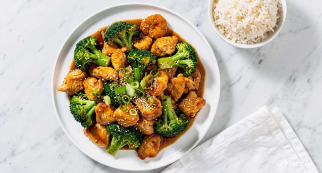 Overhead flat lay of easy chicken and broccoli stir fry on a white plate with golden chicken and bright green broccoli, garnished with sesame seeds and green onions on a white marble table