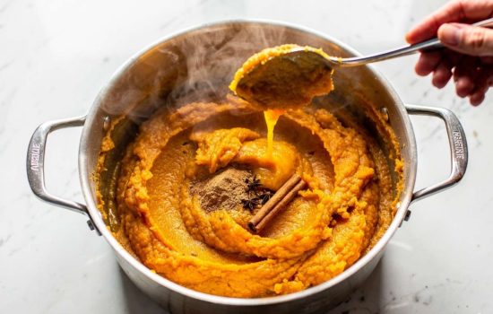 Step 2 — pumpkin puree being poured into a heavy-bottomed pot with cinnamon, nutmeg, and cloves being added, hand stirring with spoon,