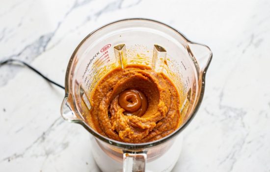 Step 3 — cooked pumpkin spice mixture being poured into a white blender, hand holding measuring cup mid-pour, orange mixture visible inside