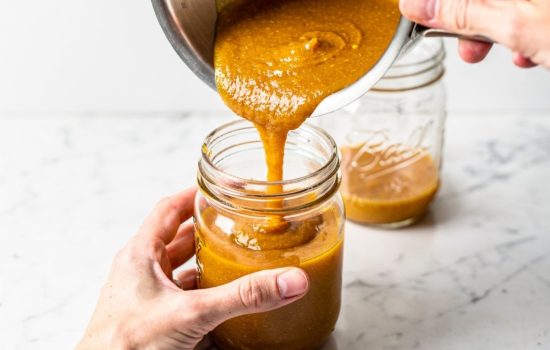 Step 5 — finished golden-orange pumpkin butter being poured from a pot into clear glass Mason jars, hands holding jar and pot, glossy butter filling jars,