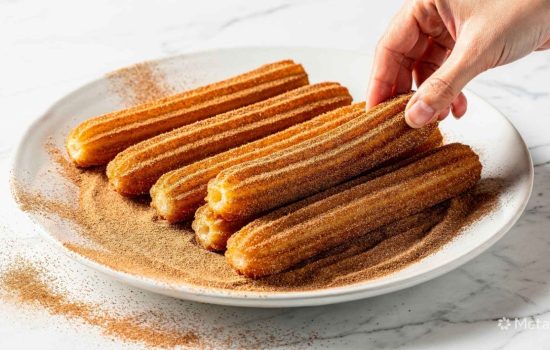 Freshly fried homemade churros being rolled in a plate of cinnamon sugar on a bright white table.