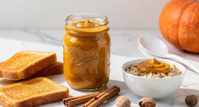 Lifestyle flat lay of pumpkin butter in a glass Mason jar on a white marble table surrounded by warm toast, oatmeal with pumpkin drizzle, cinnamon sticks, nutmeg,