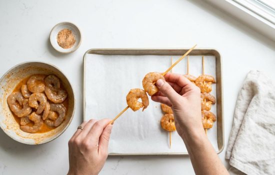 Hands threading seasoned shrimp onto skewers evenly, step 3