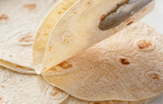 Flour tortillas warming in white skillet, tongs mid-action