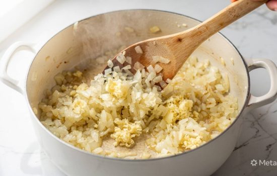 Step 2 of making Zuppa Toscana: chopped onions and minced garlic sautéing in a pot with wooden spoon mid-stir, onions becoming translucent, natural light