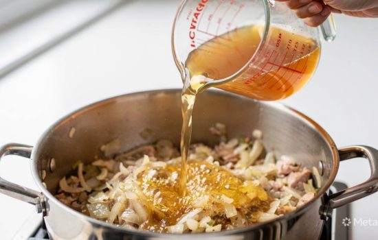 Step 3 of making Zuppa Toscana: pouring chicken broth into pot with sautéed onions and garlic, steam rising, wooden spoon visible, warm kitchen light