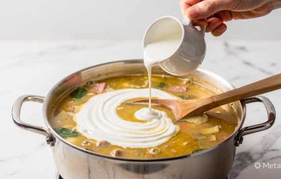 Step 5 of making Zuppa Toscana: heavy cream being stirred into simmering soup creating creamy texture, hand mid-action, soft natural light, food blog photography