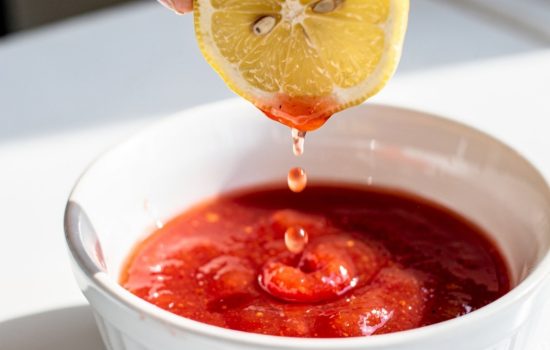 Squeezing fresh lemon juice into a bowl of red cocktail sauce on a white table.